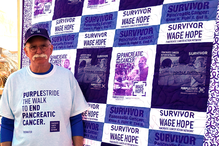 Pancreatic cancer survivor John O'Grady standing in front of a purple and white quilt he made