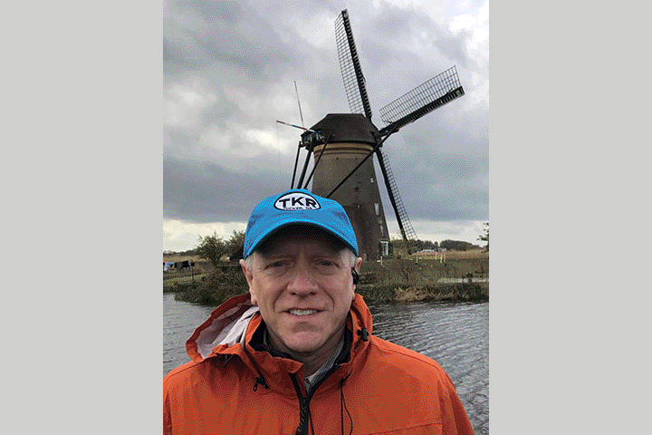 Pancreatic cancer survivor Guy Van Ort in an orange jacket and turquoise ball cap in front of a windmill