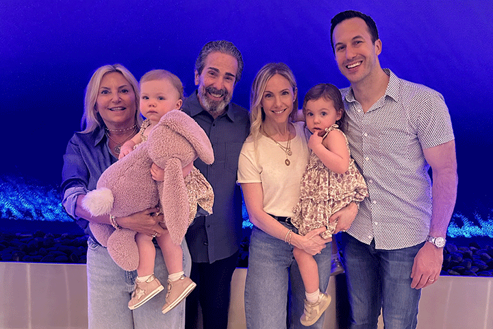 Long term survivor Susan Ganis, far left, and family. Blond woman in blue shirt and jeans holding a little girl in pink, next to a gray haired bearded man in gray and black, next to a younger blond woman in a white shirt and jeans holding another little girl, and a tall young man in a patterned shirt and jeans, against a bright blue background.