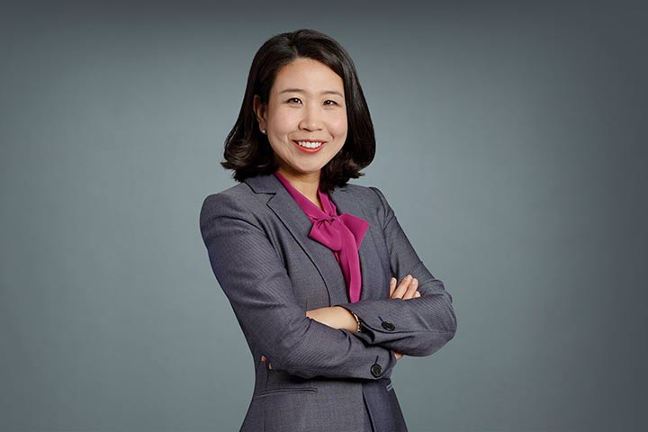 On a gray background, a woman in a pink blouse and gray jacket with dark shoulder-length hair.