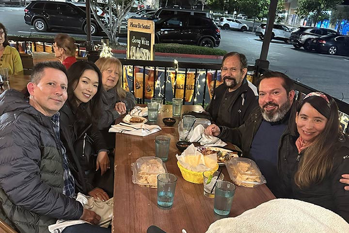 The Luna family sitting outdoors at a restaurant in cooler weather because everyone is wearing jackets, with her son, a woman, Roberta (a blonde woman), her husband (an older man), her other son, and a young woman.
