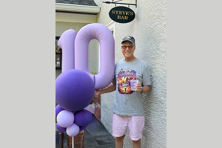 Man in shorts, a t shirt and a ball cap next to purple balloons with the number 10