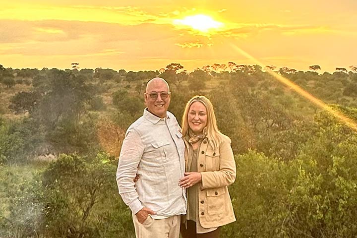 A bald man with glasses and a blonde woman stand on an overlook with the sun setting behind them