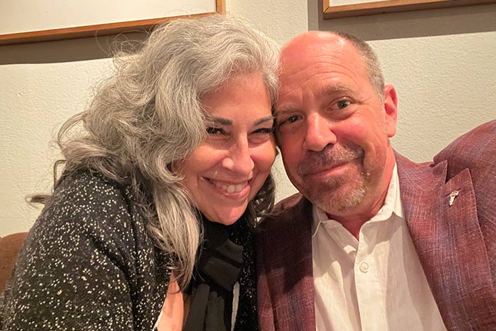 Smiling woman with long gray hair with her head next to a balding man with a trimmed beard