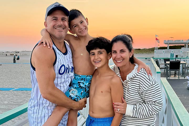 A family--Husband in a ball cap and tank top, two brown haired sons in swim trunks, and a wife with brown hair in a beach coverup--standing on a boardwalk with the sunset in the background