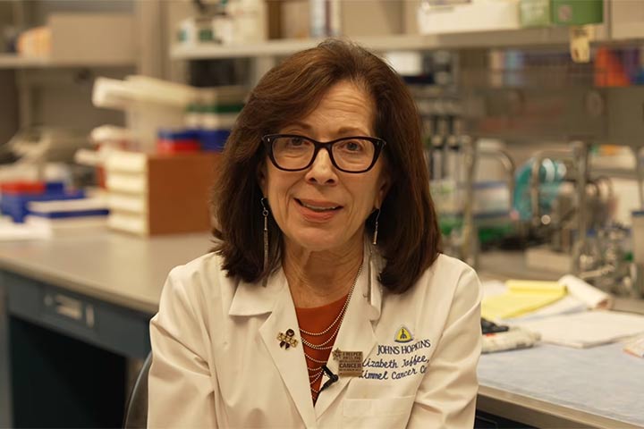 Woman in a science research lab in a white coat, with brown hair and glasses.