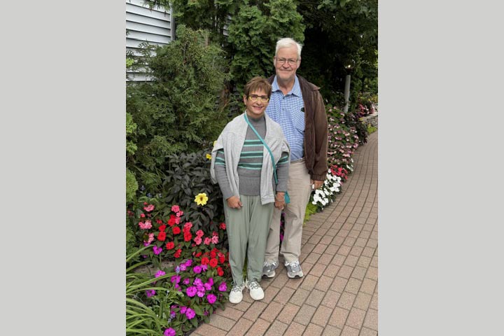 An older couple standing outside with bright pink and red flowers nearby