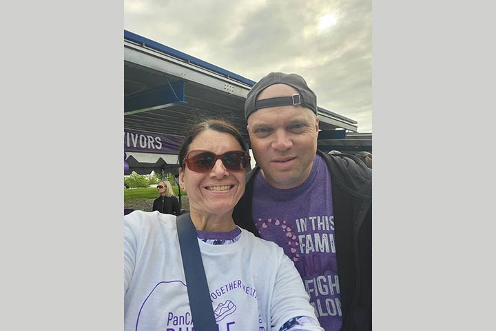 Woman in sunglasses, a white shirt and crossbody bag with a man in a reverse ball cap, purple shirt and sweatshirt
